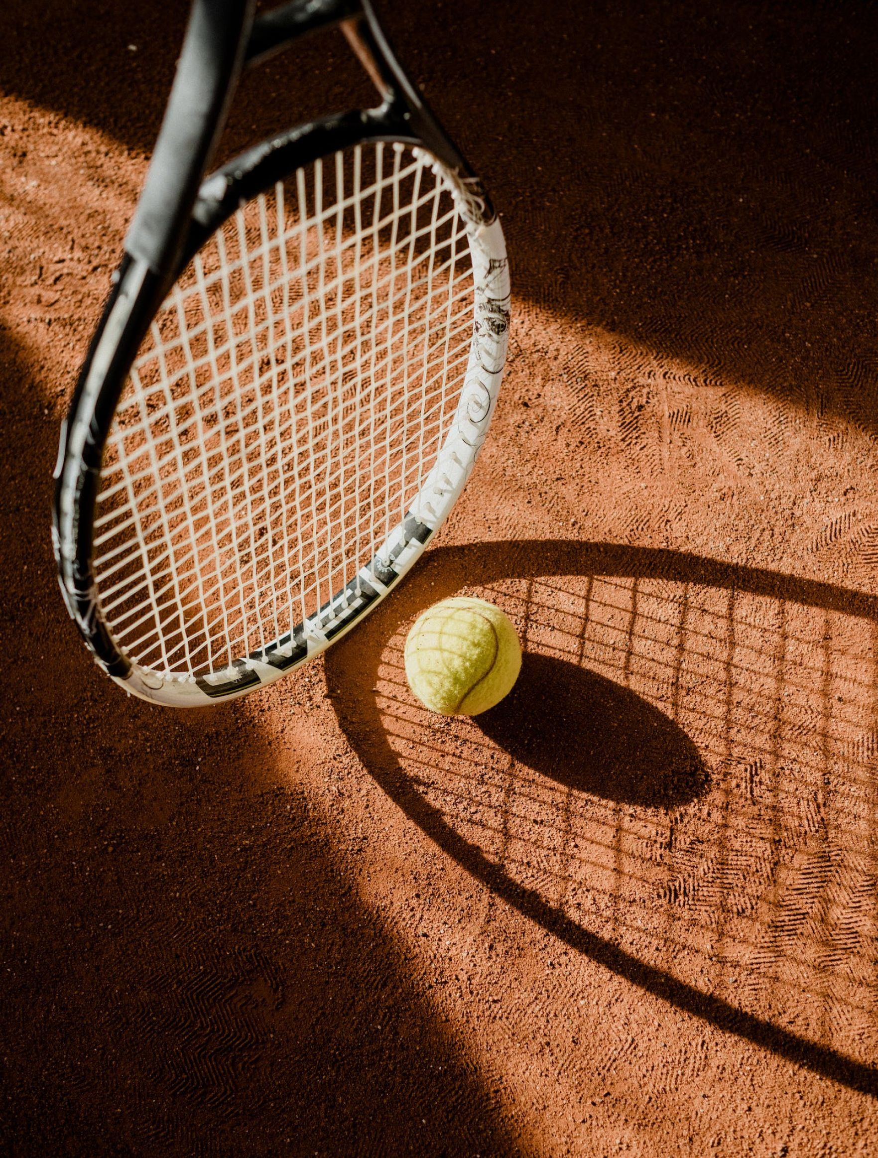 Tennis racket and ball on a clay court, with a long shadow in warm sunlight