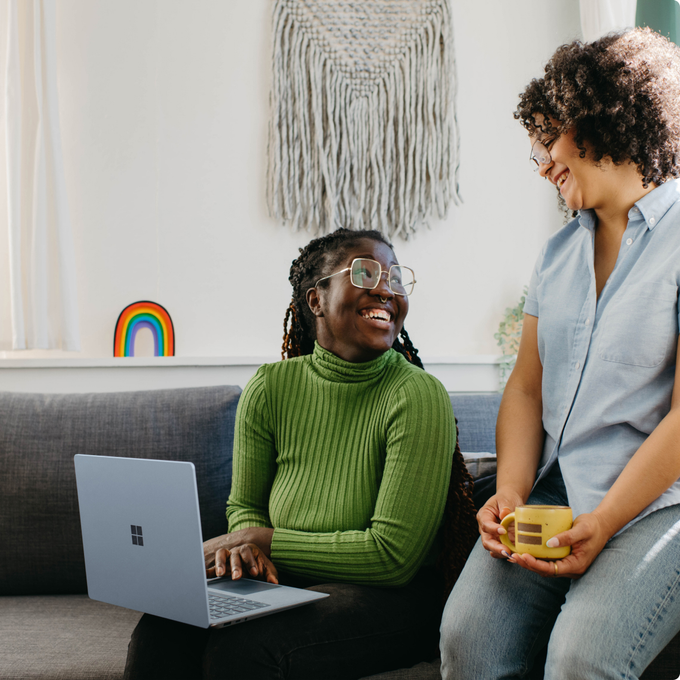 Two women are sitting on a couch with a laptop and a cup of coffee