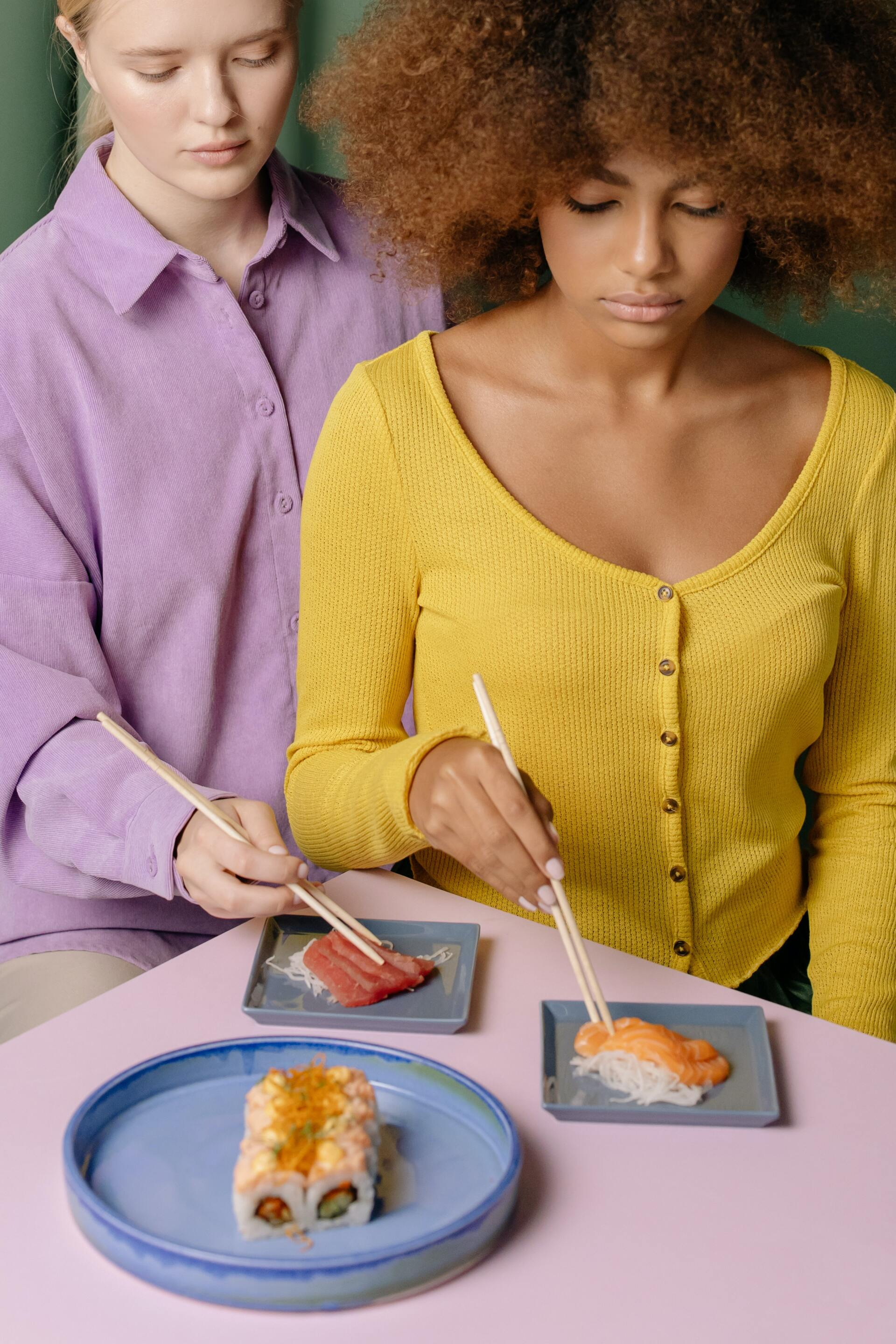 Two people eating sushi at a pink table. One in a yellow shirt, one in a purple shirt.
