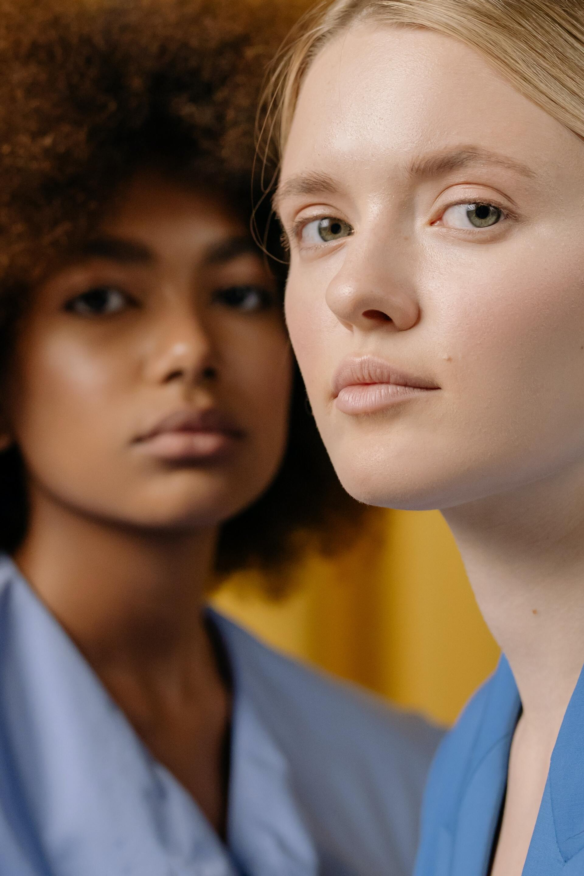 Two women in blue scrubs, one with curly hair in the background and one with blond hair in the foreground, looking forward.
