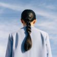 Person seen from behind with long dark braid wearing a light blue shirt against a plain blue background