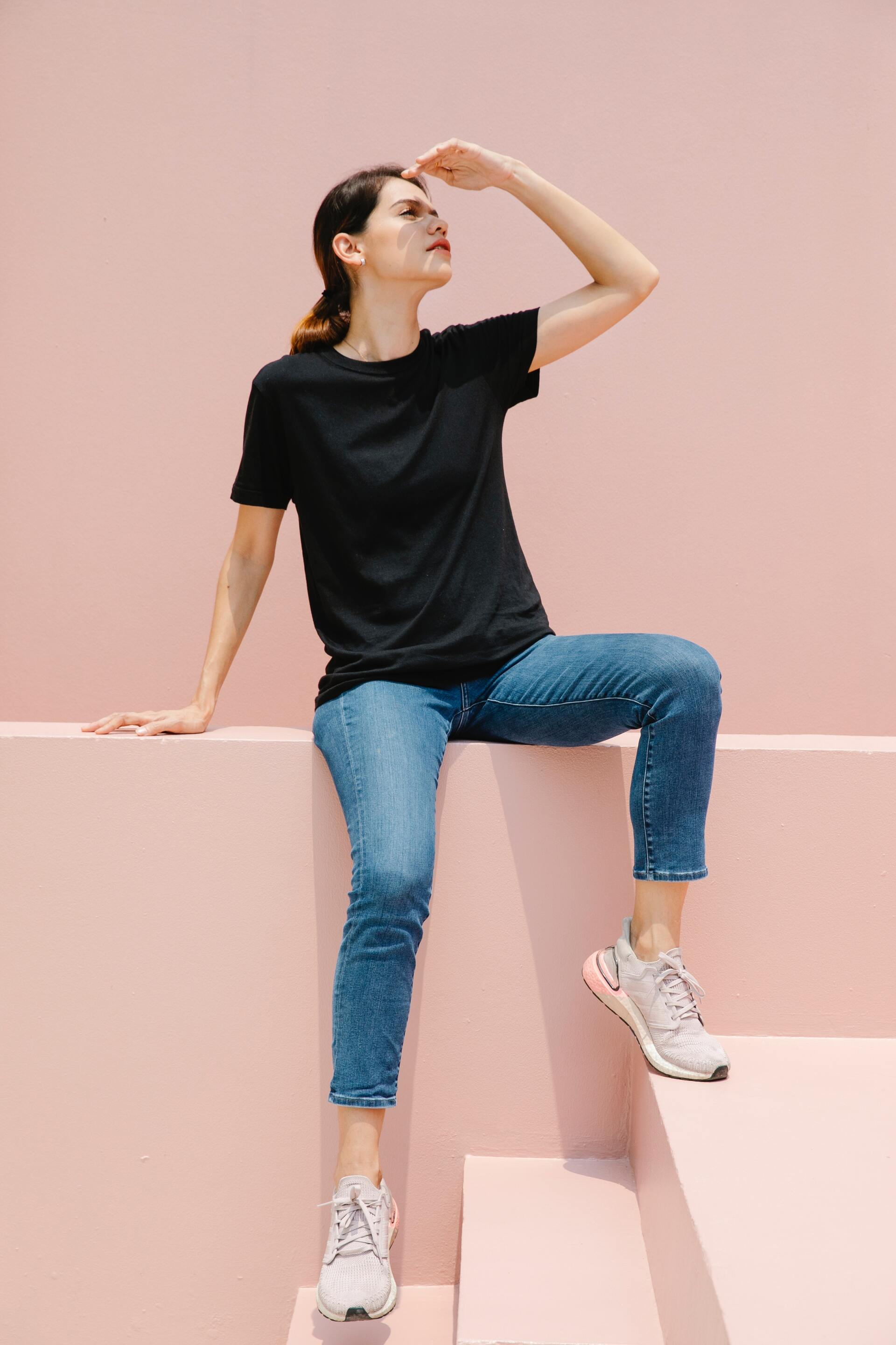 Una mujer con una camiseta negra y jeans está sentada en una pared rosa.