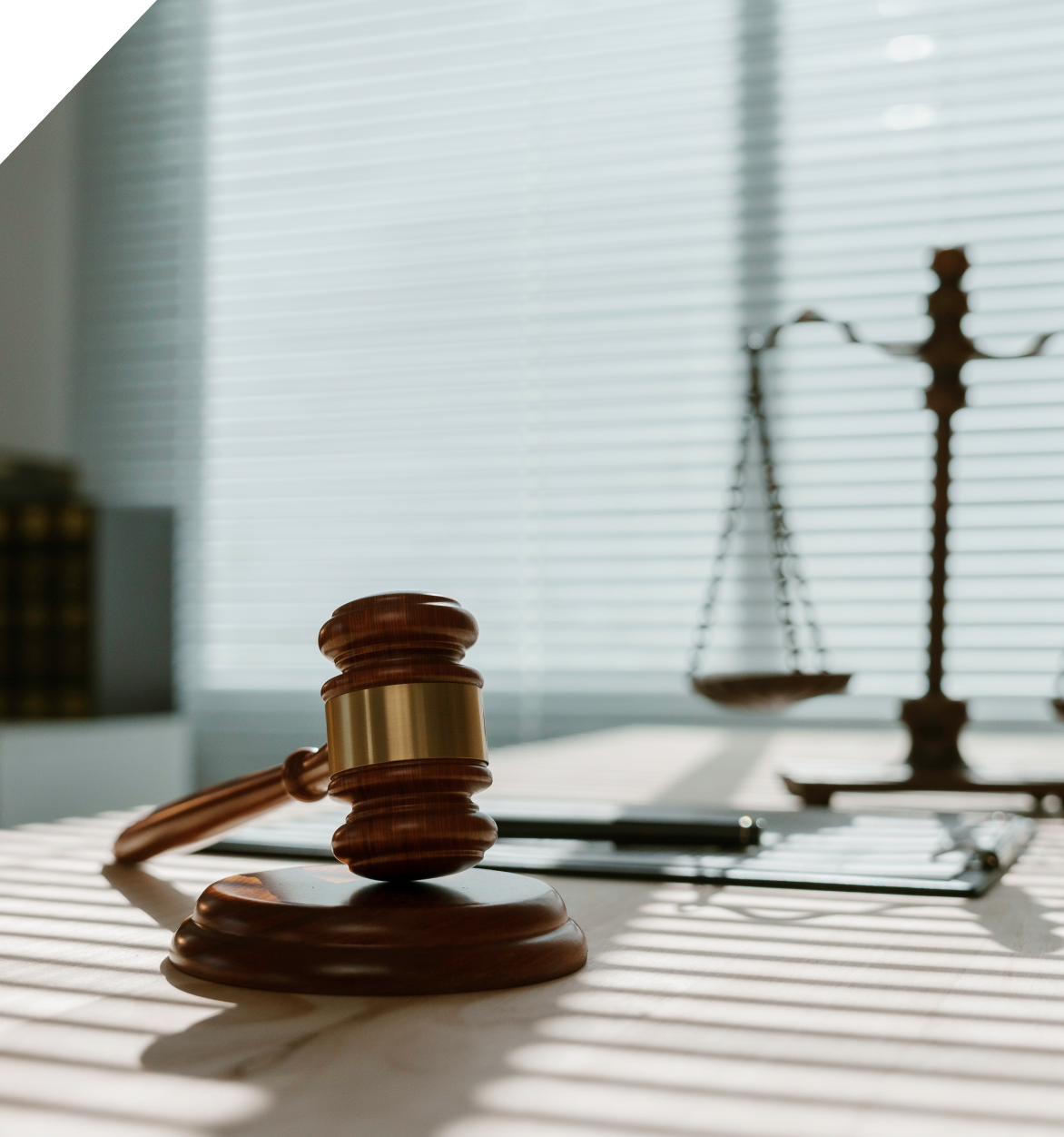 Su una scrivania, sullo sfondo di persiane, si trovano un martelletto da giudice in legno e una bilancia metallica.