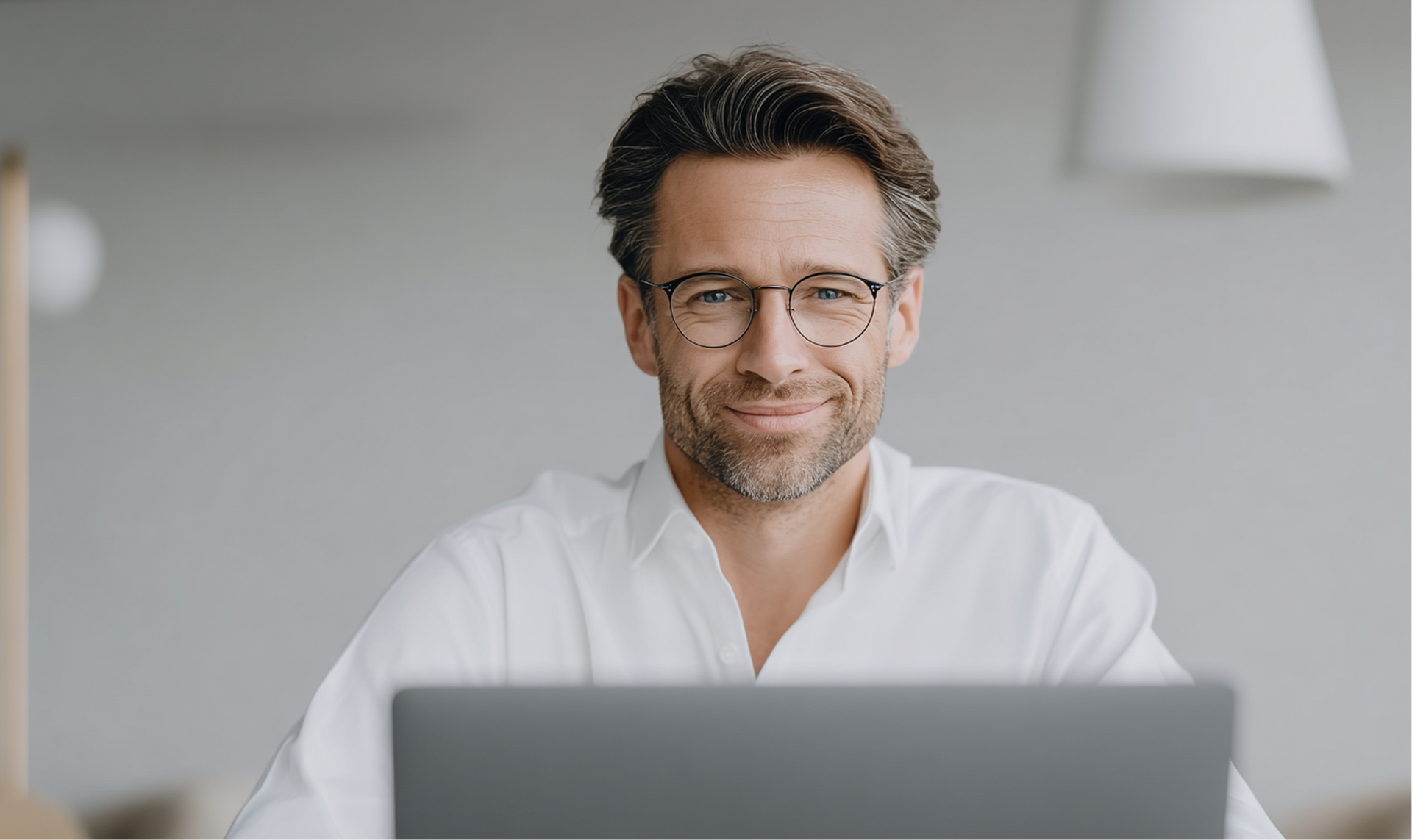 Un uomo con gli occhiali sorride alla telecamera mentre lavora al computer portatile. Indossa una camicia bianca in un ufficio luminoso.
