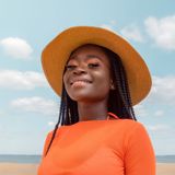 A woman wearing a straw hat and an orange shirt is smiling on the beach.