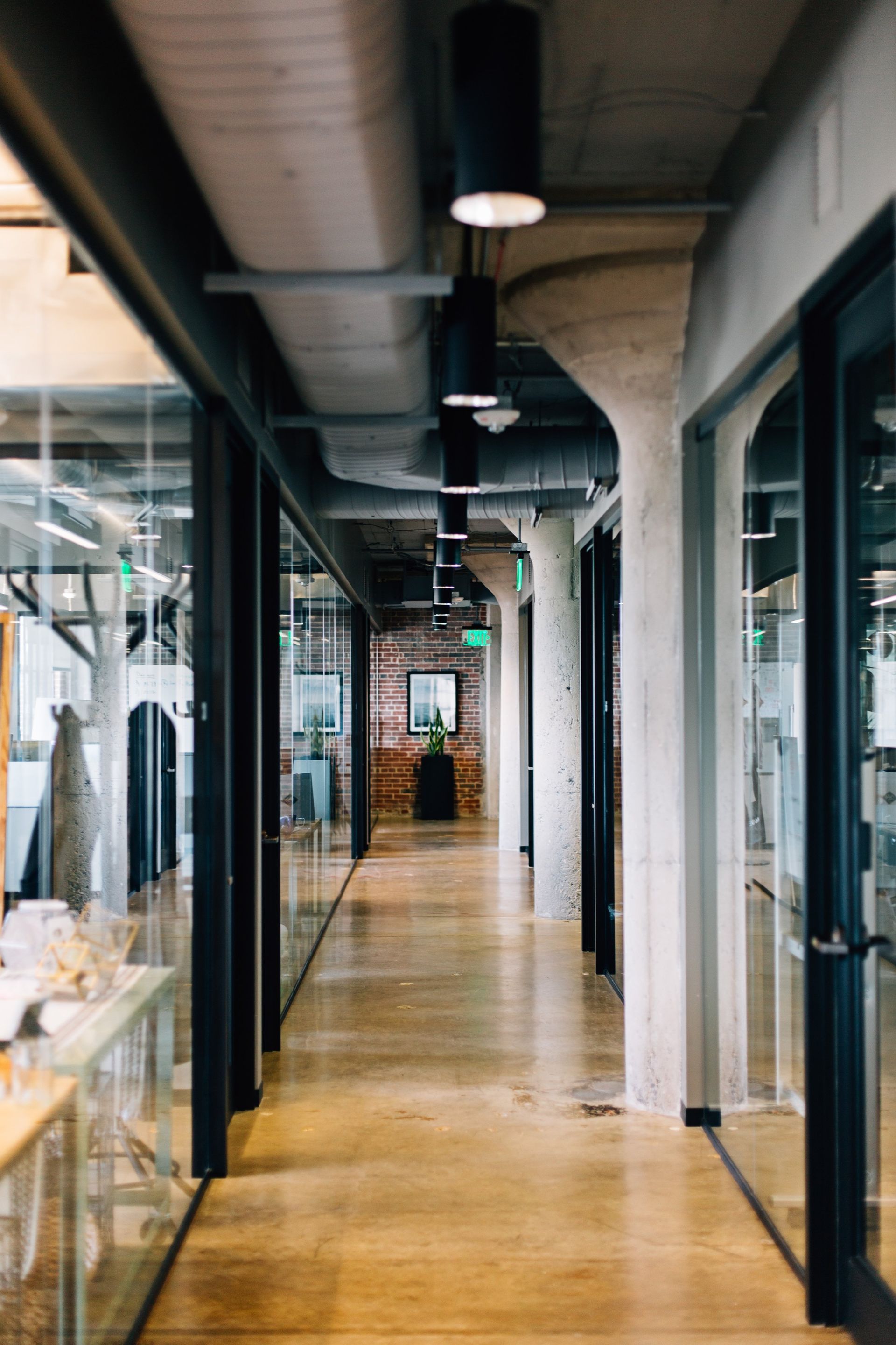 Long hallway in an office with glass-walled rooms, concrete columns, and overhead lighting.