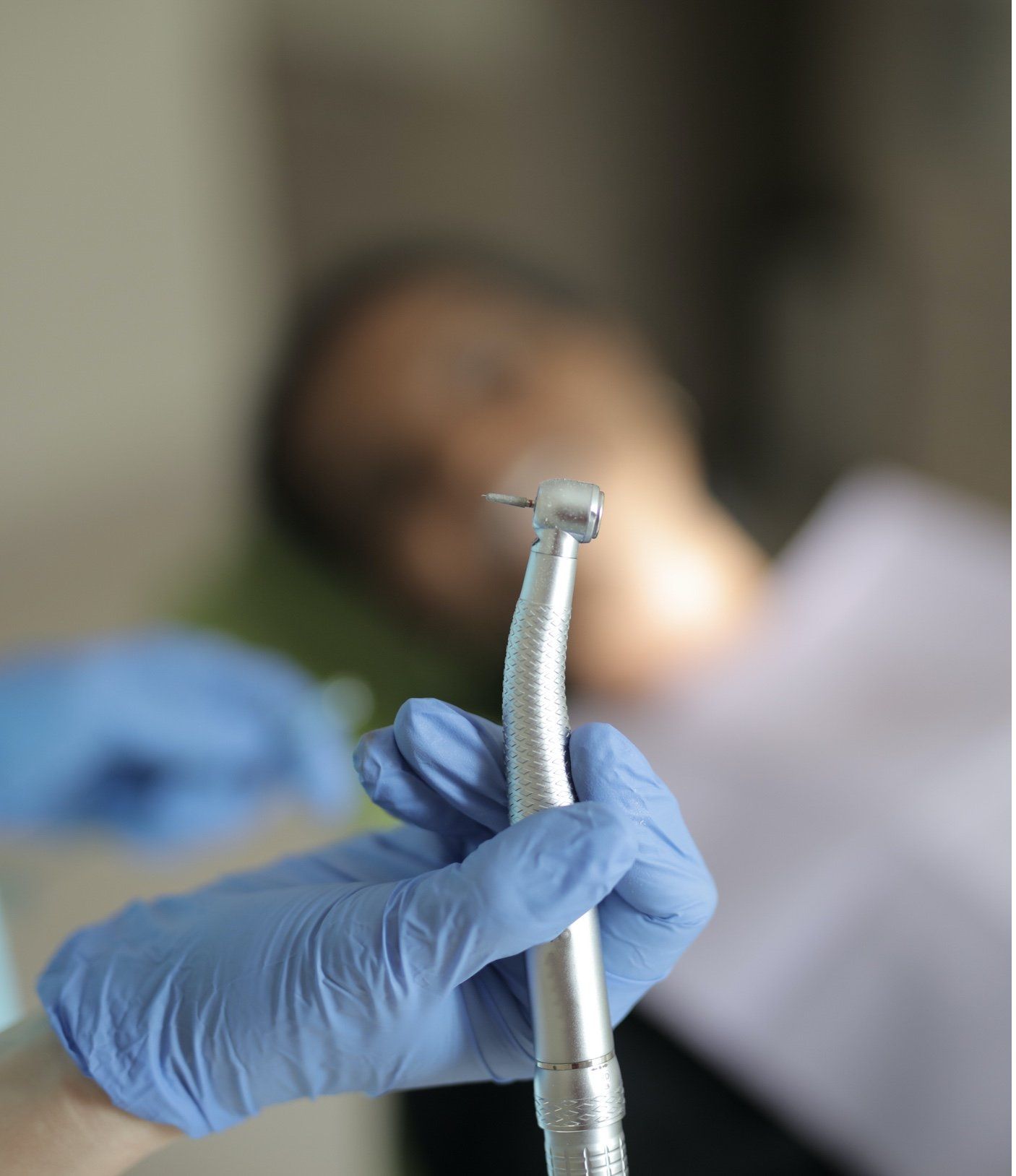 Mano de dentista en guante azul sosteniendo un taladro dental, paciente en el fondo.