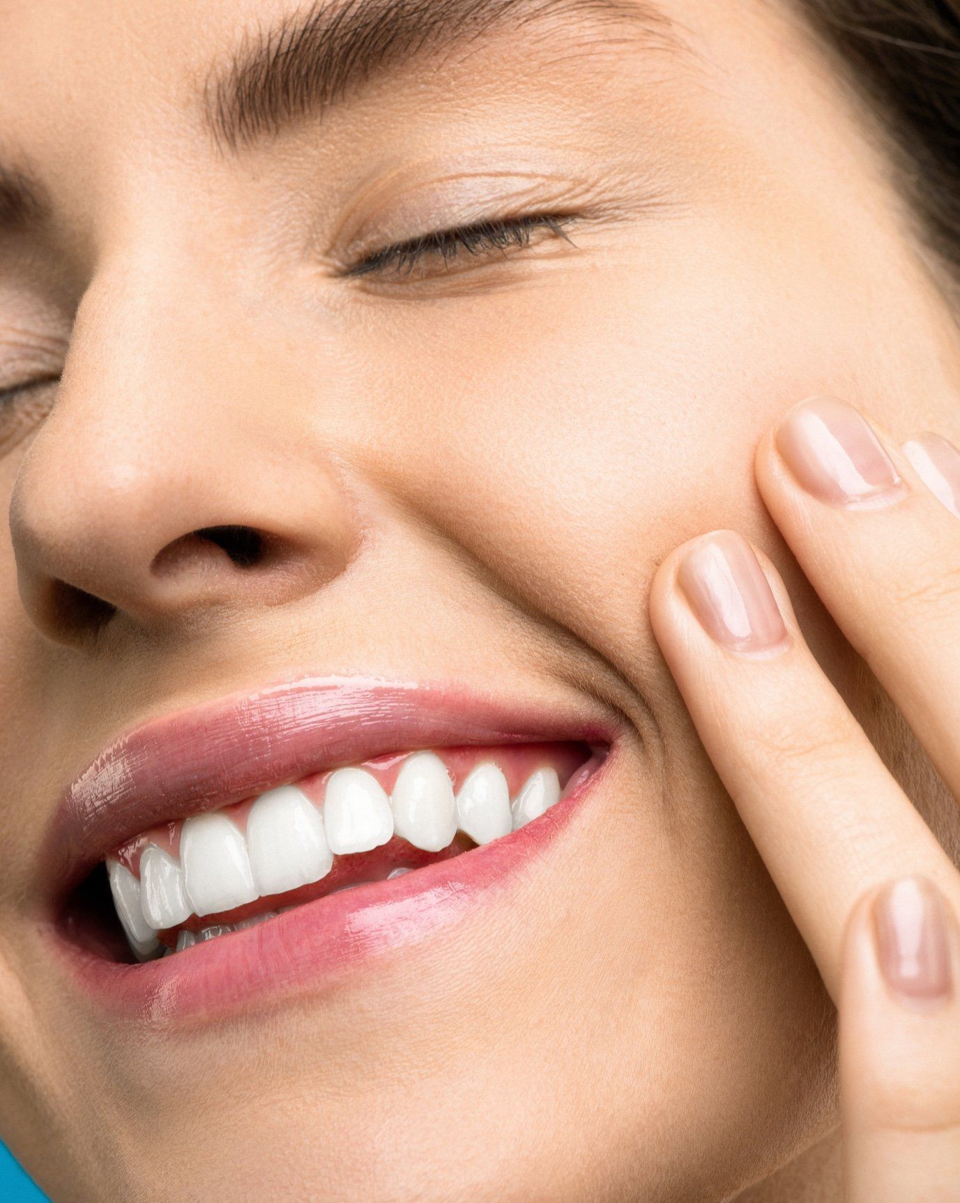 Mujer con los ojos cerrados, sonriendo, tocándose la mejilla. Lápiz labial rosa, piel clara y uñas naturales.