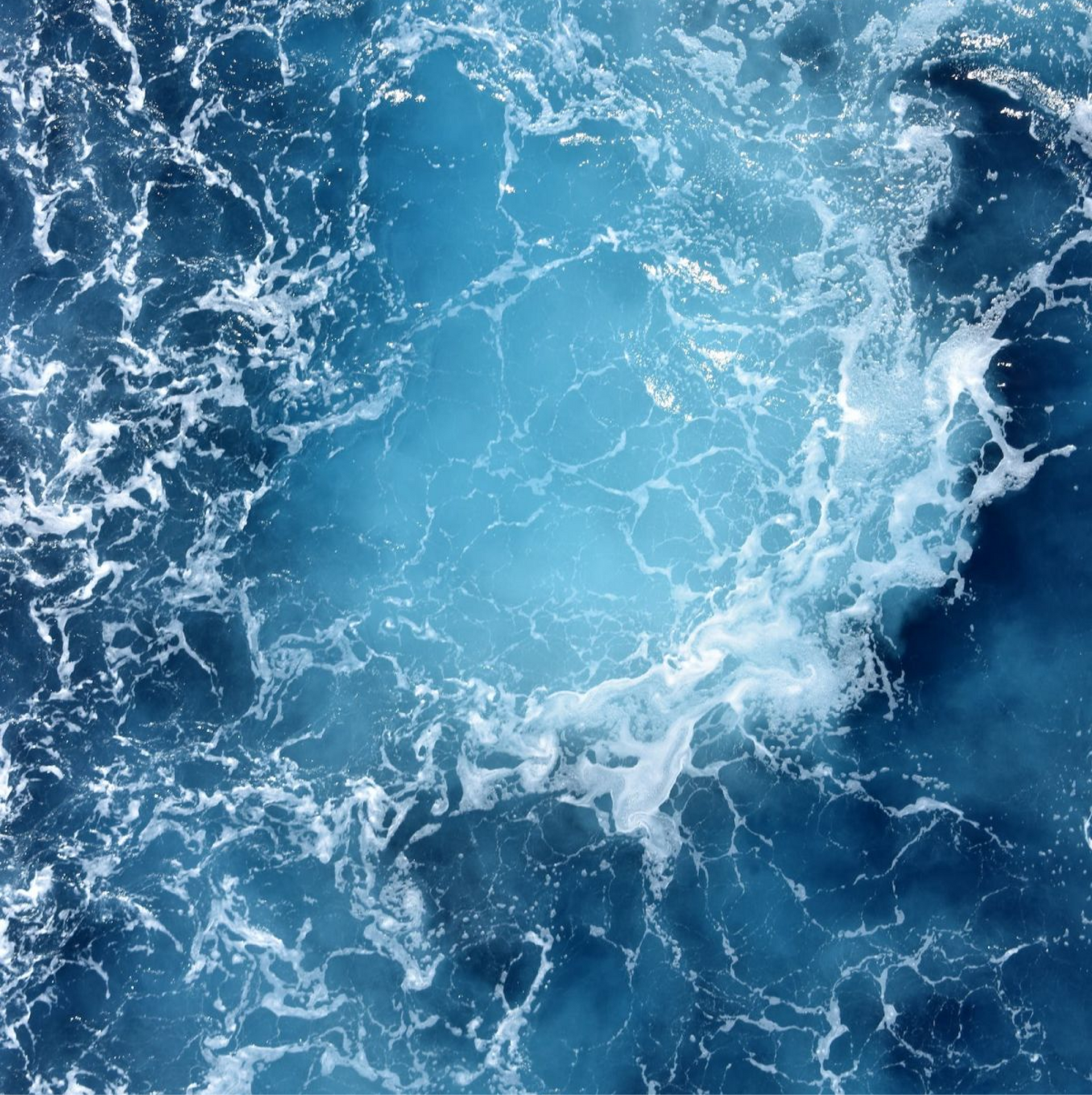 Veduta aerea di acque oceaniche di un blu intenso con schiuma bianca e vorticosa che crea un disegno astratto.