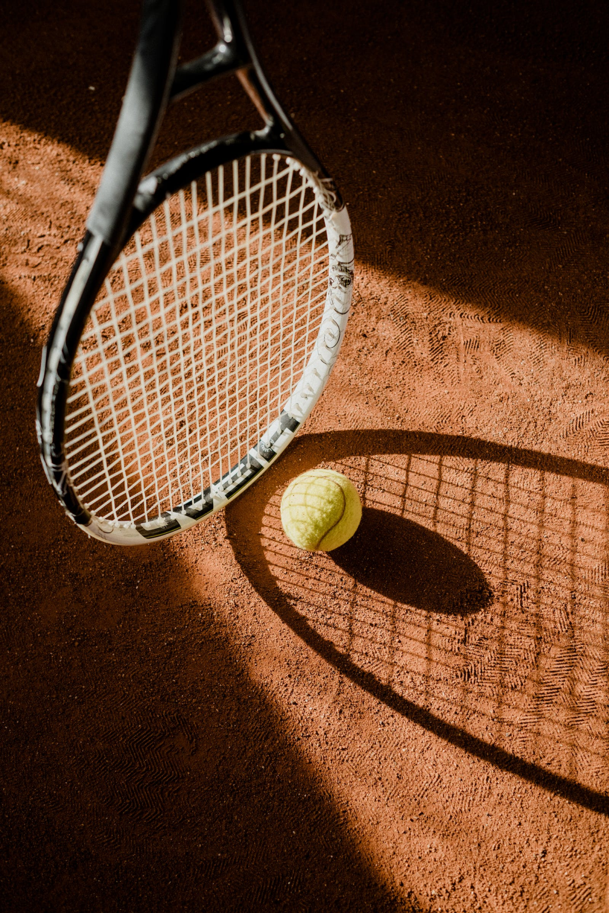 Tennis racket and ball on a clay court, casting a long shadow.