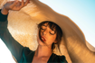 Woman with freckles and bangs shielding face with large straw hat against blue sky.