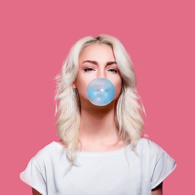 A woman is blowing a bubble of gum in front of a pink background.