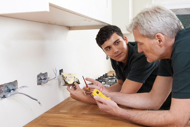 Two men are working on an electrical outlet in a kitchen.