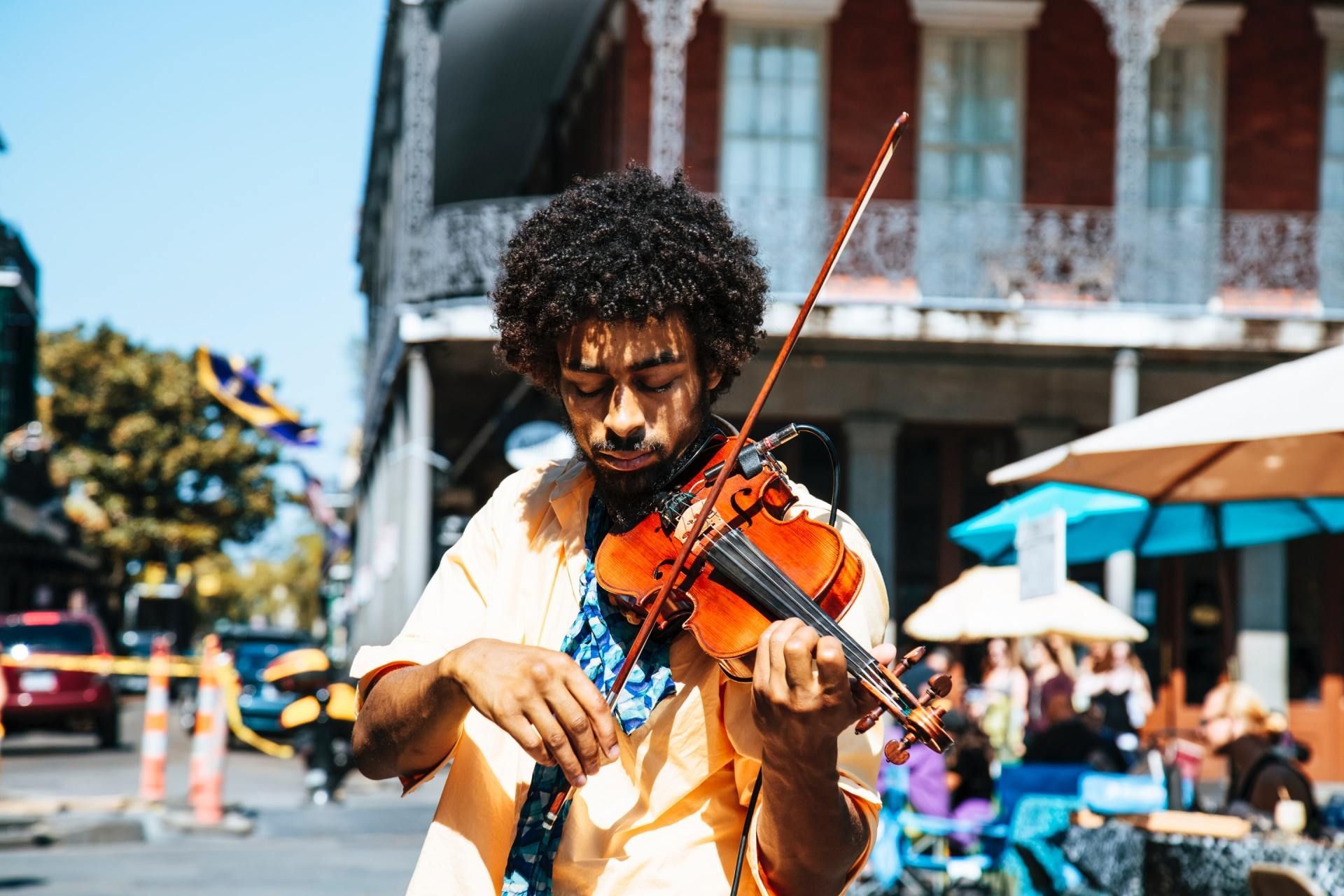A Black musician with curly hair plays the violin on a street corner.