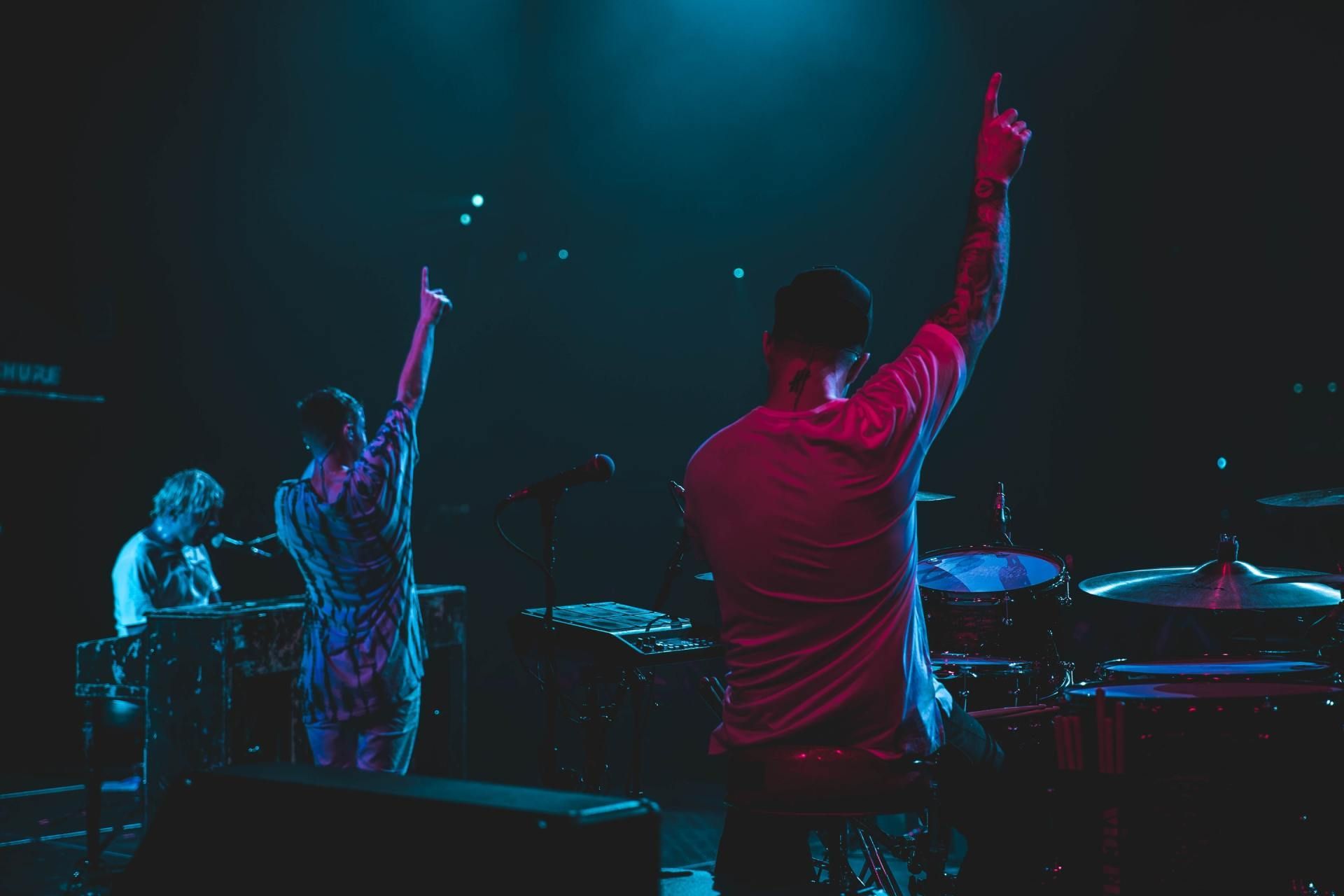 Two band members on stage with arms raised in front of a drummer and keyboardist, lit by blue and red lights.