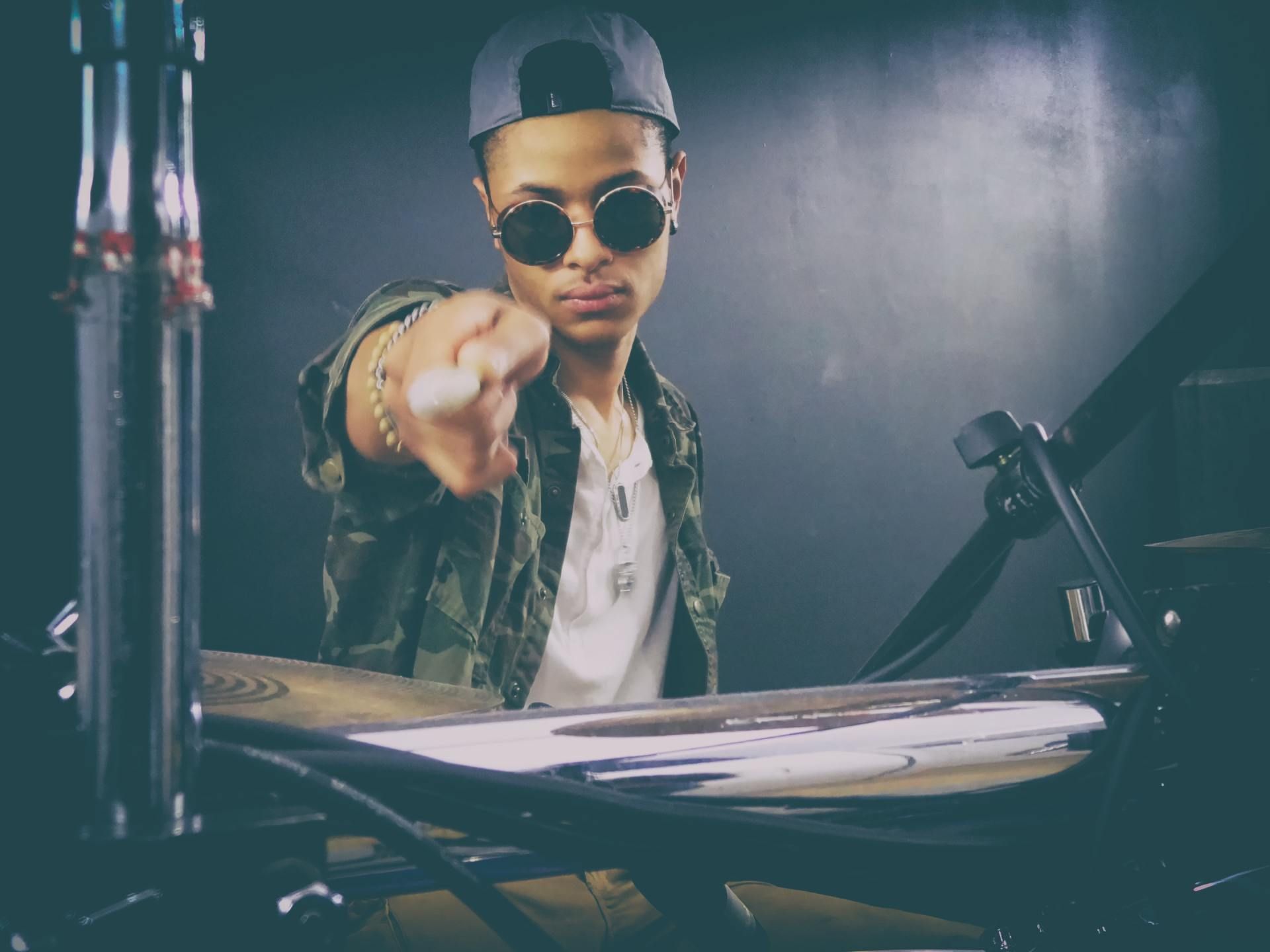Person with sunglasses and cap points towards camera, sitting at a drum set in front of a dark background.