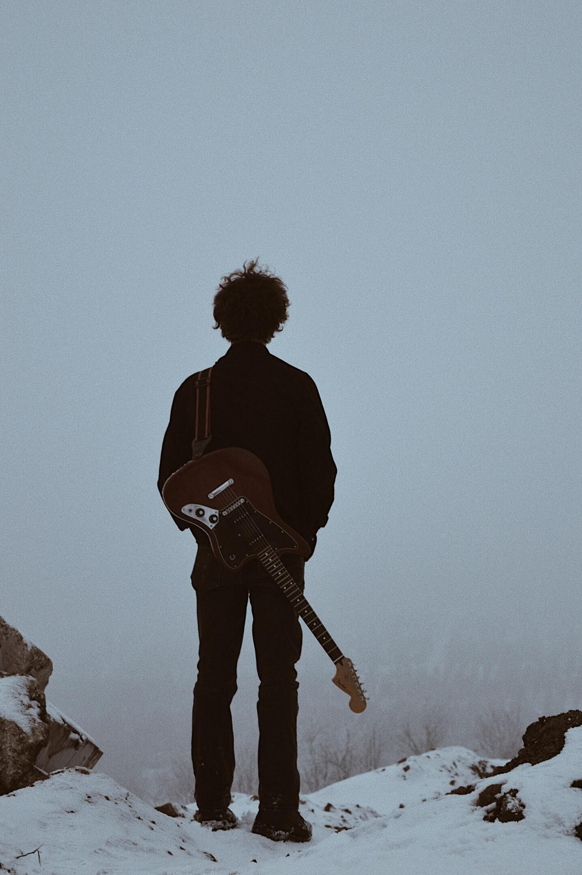 Person with guitar, standing on snowy cliff, looking out at a foggy landscape.