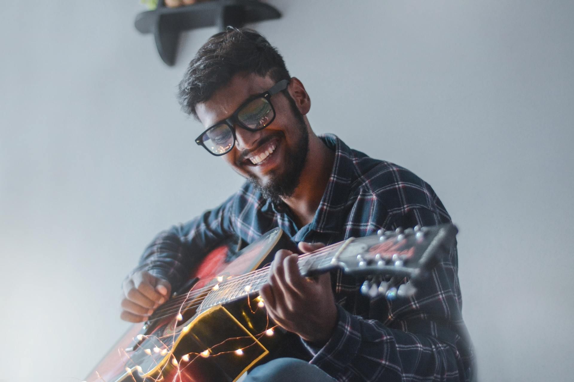 Man with glasses happily playing guitar with string lights.