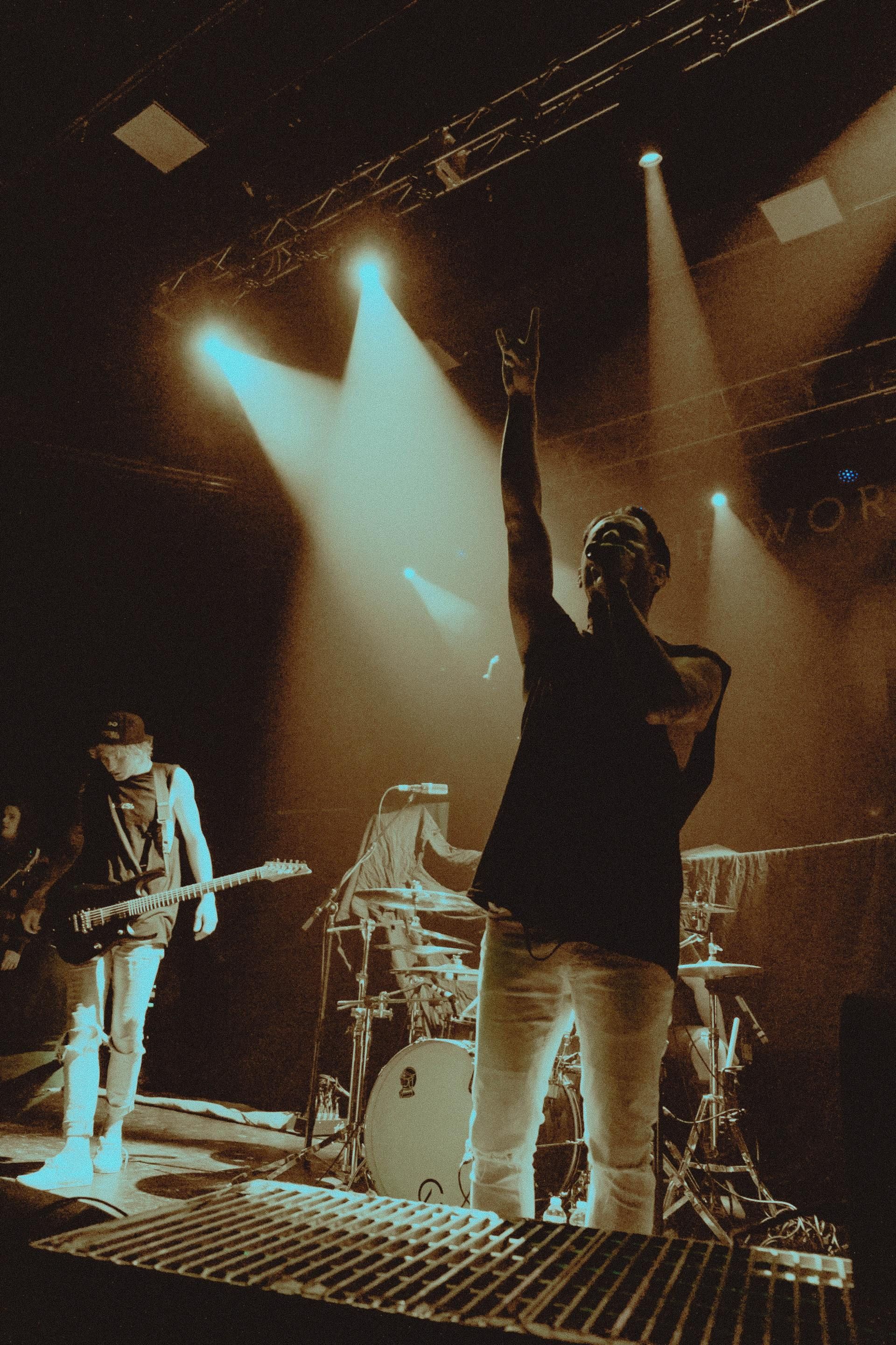 Band performing on stage under bright spotlights. Singer with arm raised, guitarist plays.
