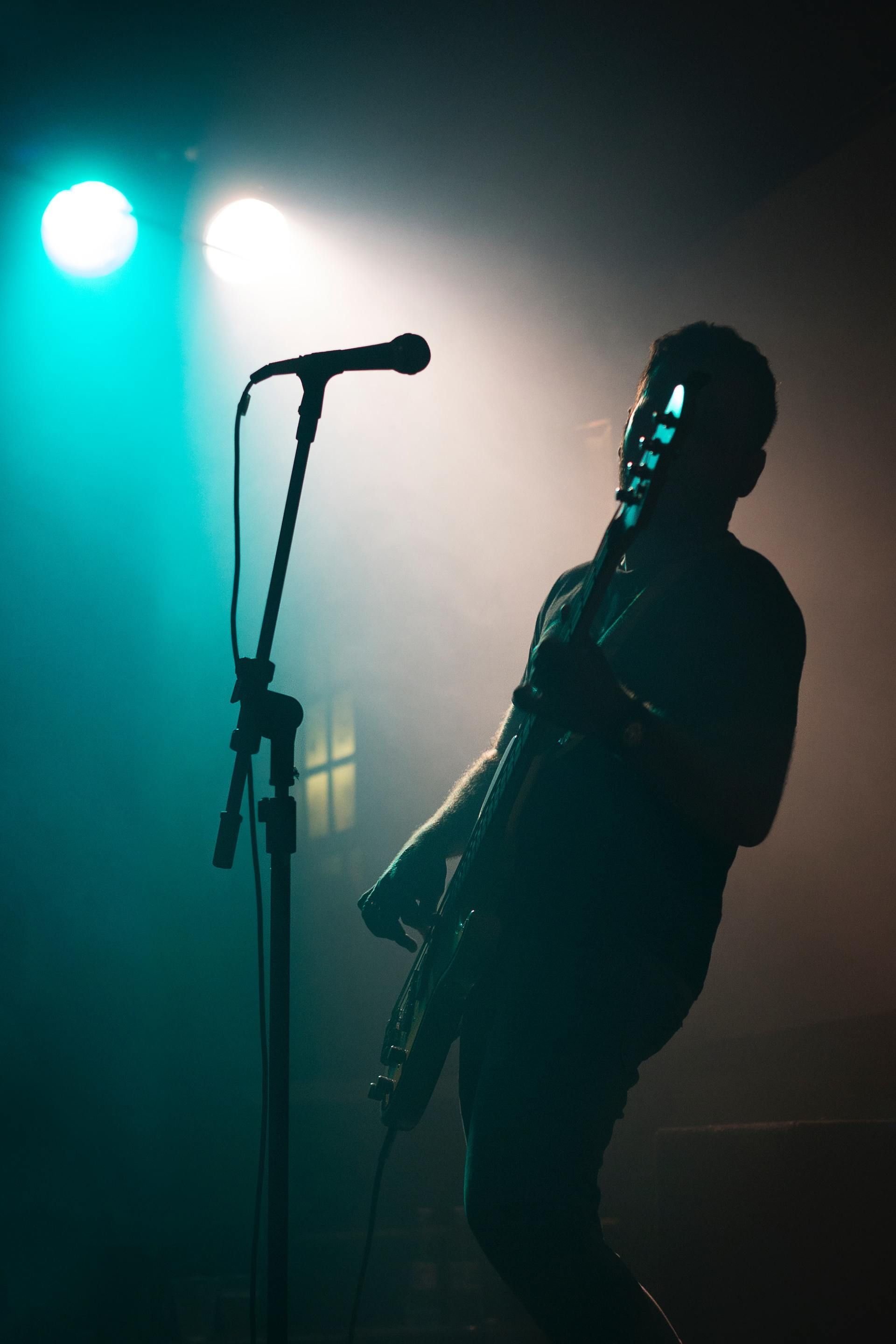 Silhouette of a musician on stage, holding a guitar under teal and orange stage lights.