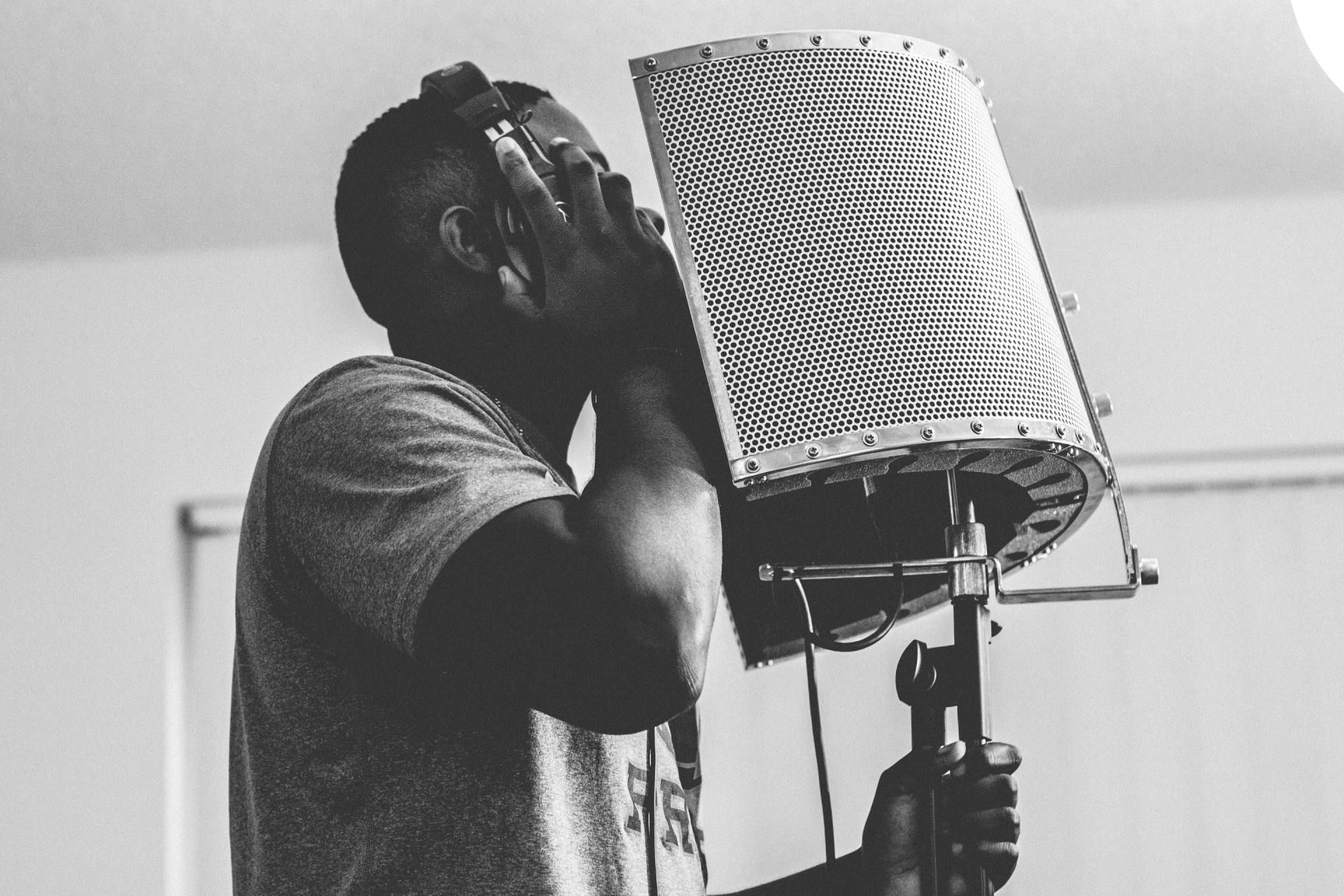 Black and white photo of a man recording vocals into a microphone in a studio setting.