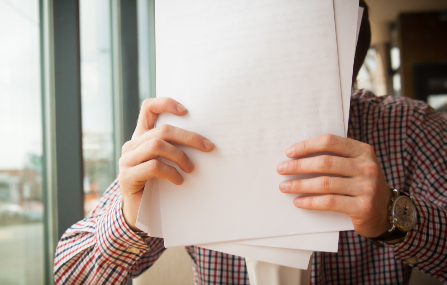 Personne tenant des feuilles blanches vierges devant son visage, chemise à carreaux rouges, montre au poignet, fenêtre floue en arrière-plan.