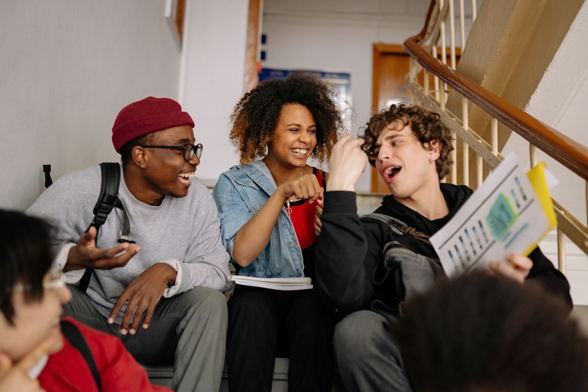 Group of students laughing on stairs, holding papers; one with curly hair points, another wearing a burgundy beanie.