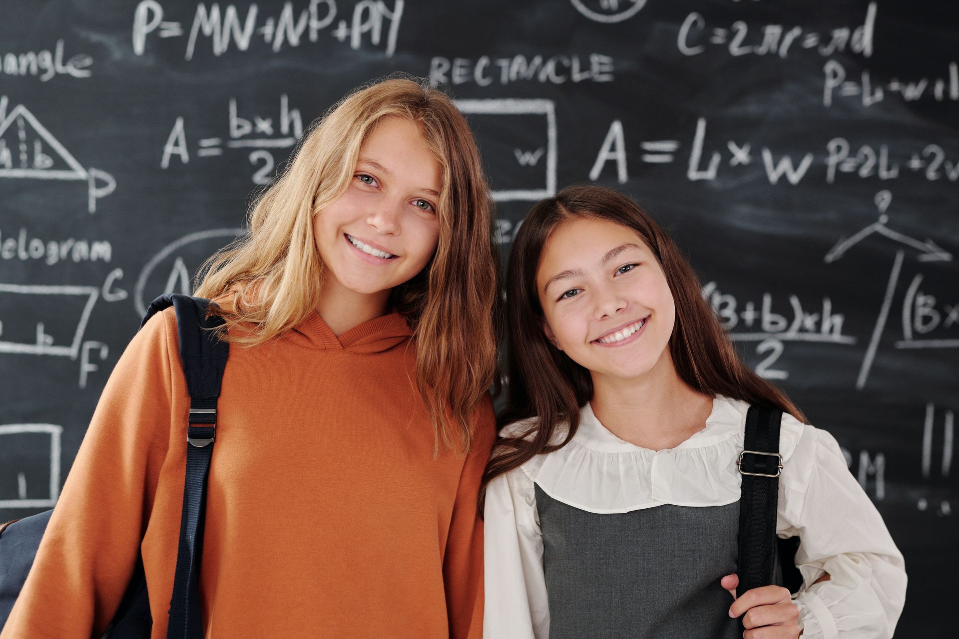 Two smiling girls in front of a chalkboard filled with math formulas.