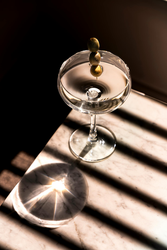 Cocktail in a martini glass, garnished with olives. Sunlight casts shadows across a marble surface.