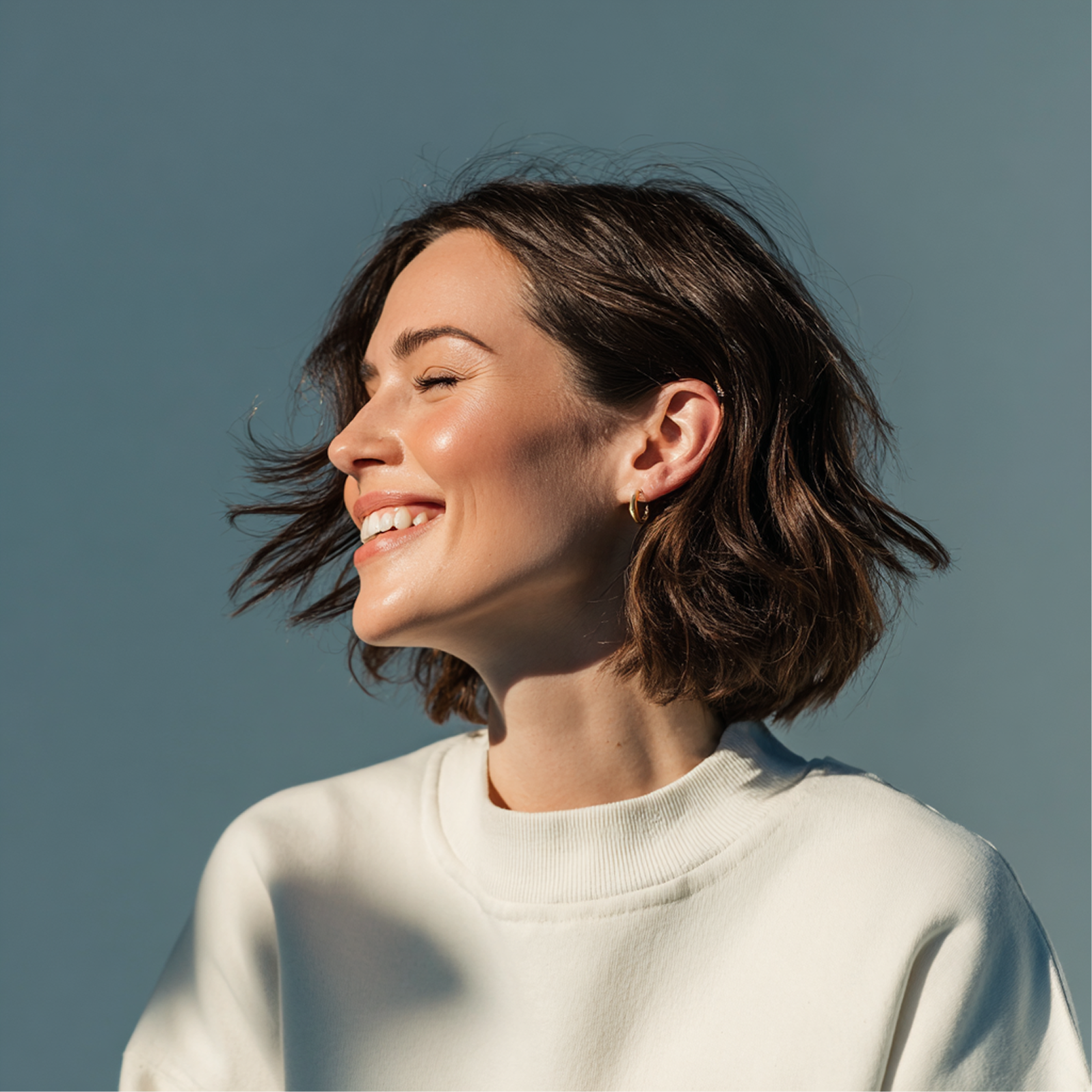 Woman smiles, eyes closed, head tilted toward sunlight. Short brown hair, white sweater. Blue background.