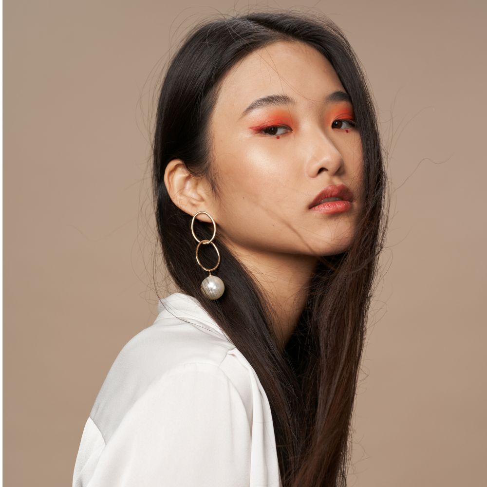 Woman with long black hair, wearing gold earrings and orange eye makeup, facing right.