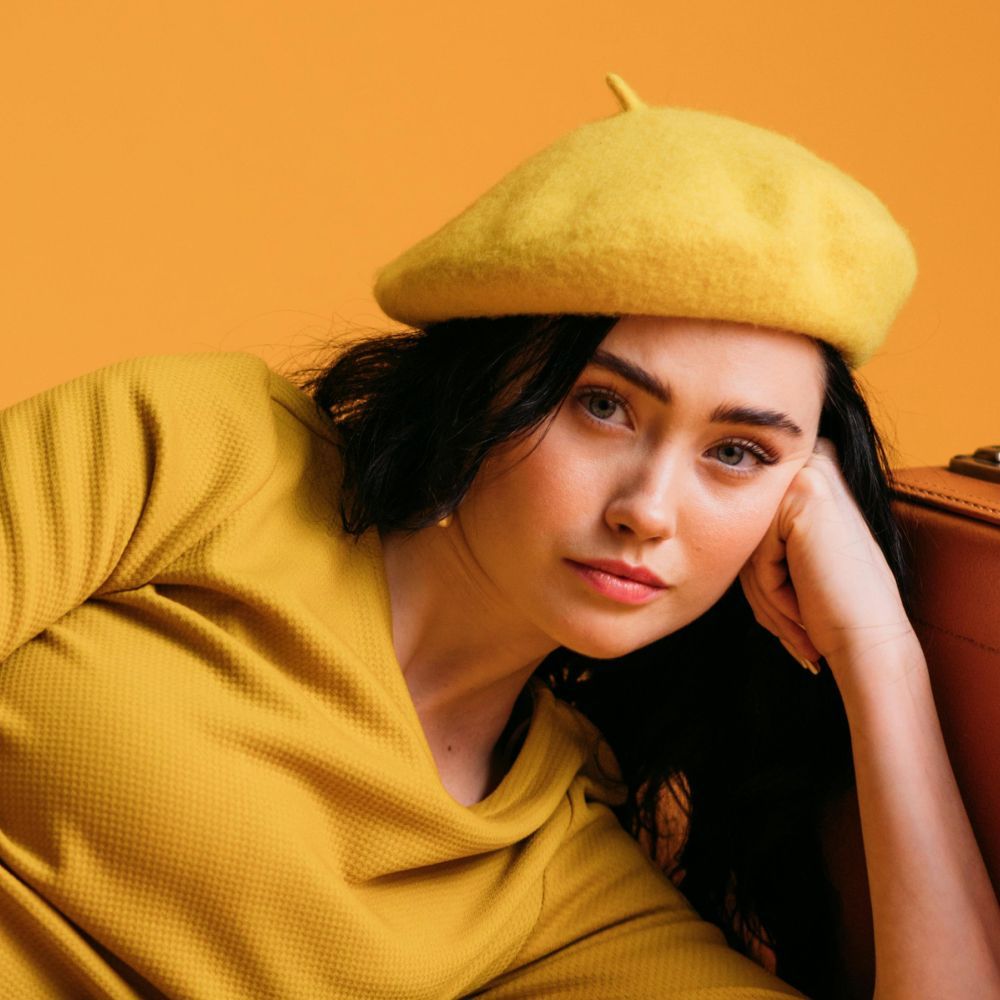 Woman in yellow beret and shirt, resting head on hand, posing next to a brown suitcase against a yellow background.