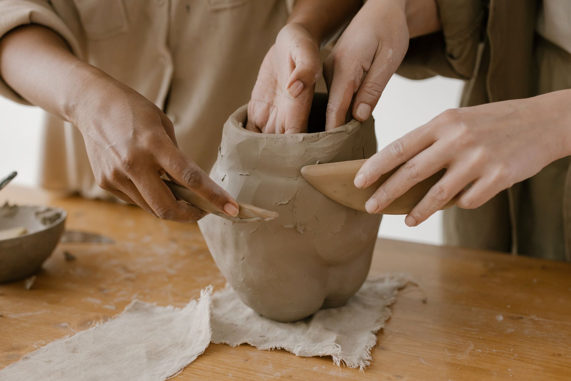 Two pairs of hands smooth and shape a raw clay vessel sitting on a wooden table.