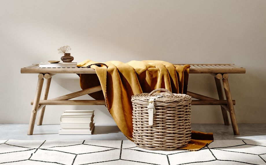 A wooden bench holds a mustard-colored throw, a small vase, and books, with a wicker basket beneath, on a patterned rug.