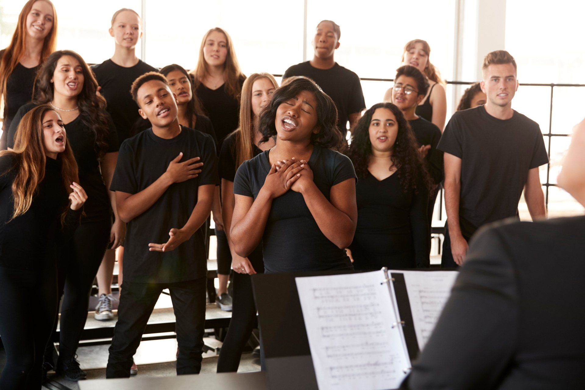 Choir in black shirts singing, led by a conductor with sheet music in a brightly lit room.