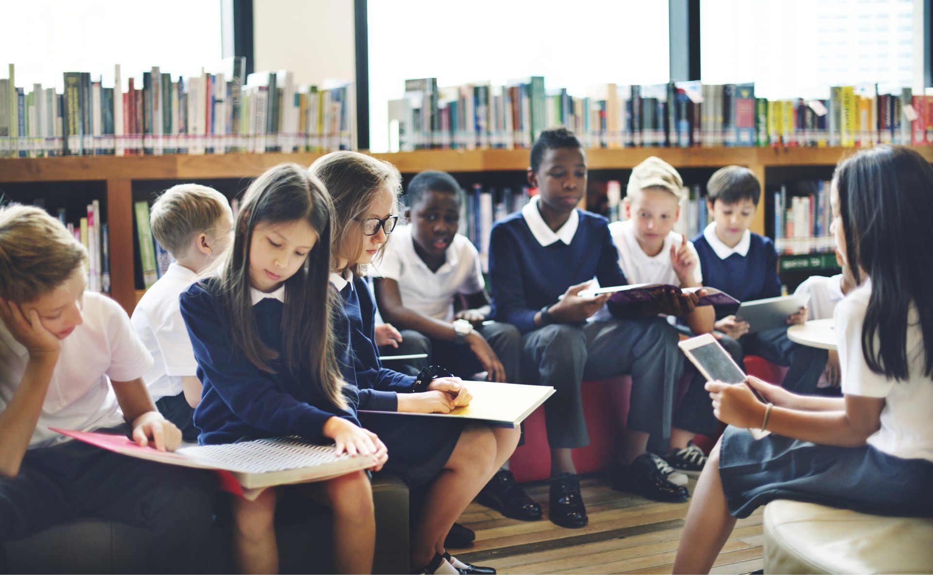 Children in school uniforms gathered in a library, some reading books, others looking at a tablet.
