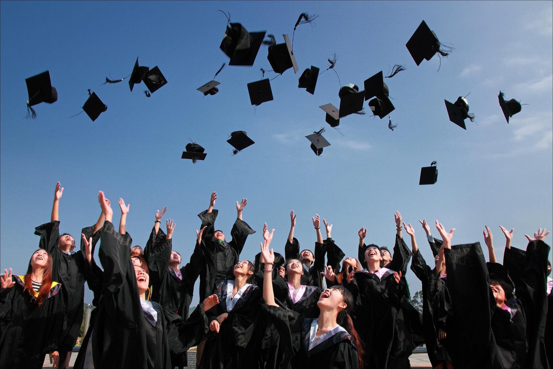 Graduates in black gowns and caps throwing their mortarboards in the air against a bright blue sky.