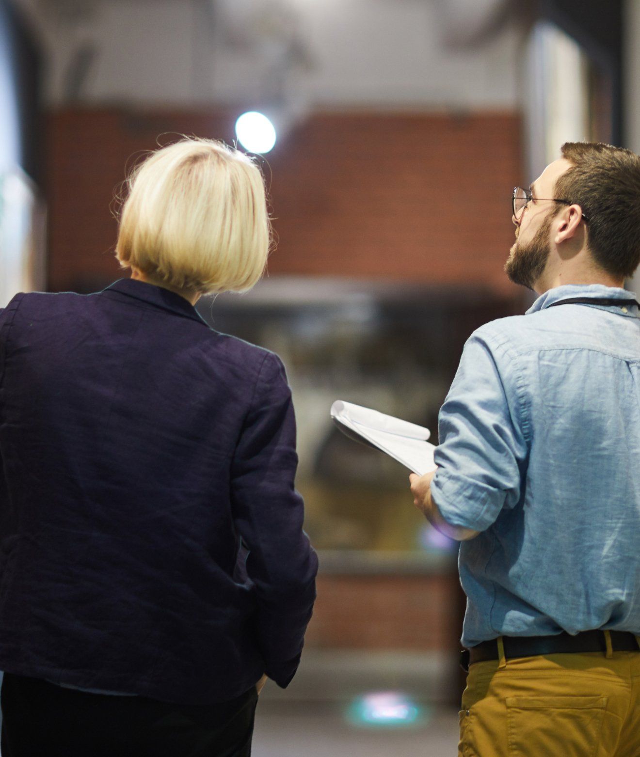Two people looking up in a hallway; a woman in a blazer and a man in a denim shirt holding papers