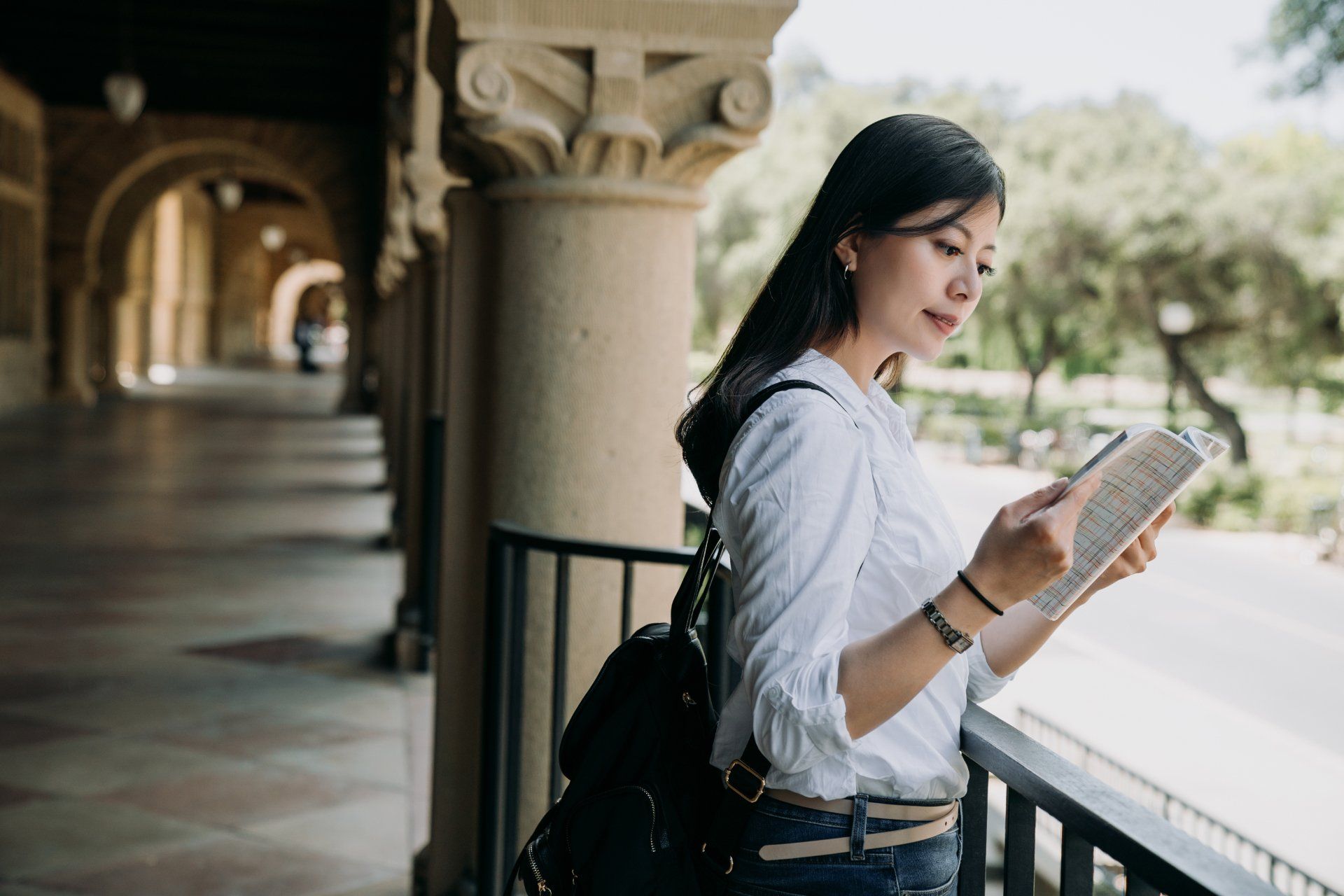 A young Asian woman reads a book on a balcony with stone columns in a university setting. 