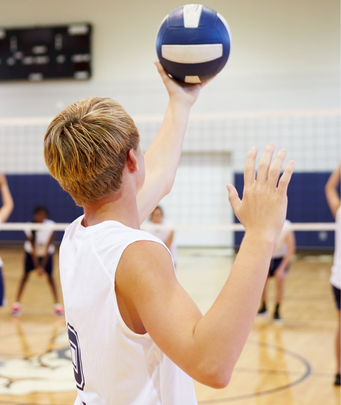 Blond volleyball player prepares to serve, ball overhead, other players in the background. Indoors, volleyball court.