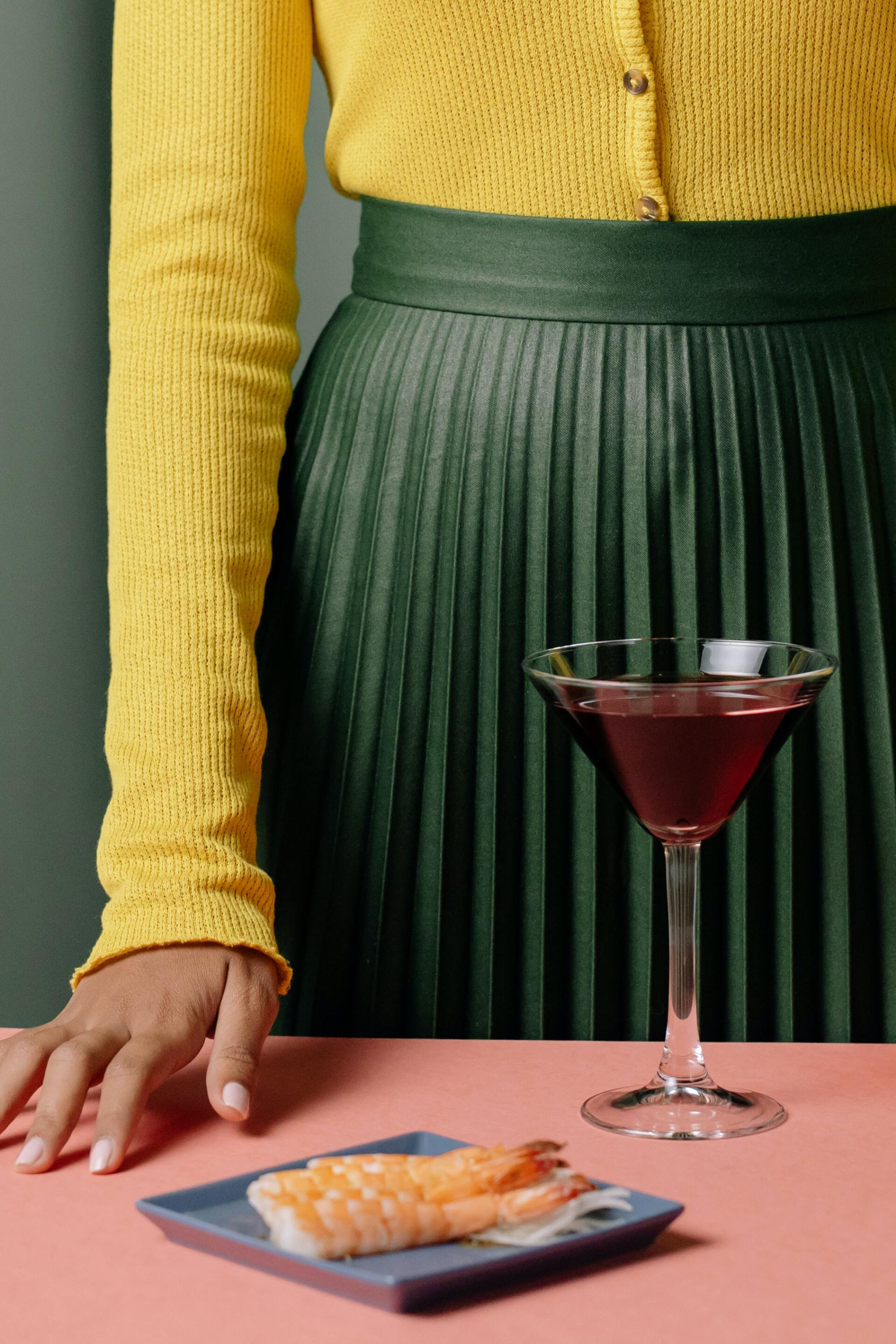 A woman in a yellow sweater is standing next to a martini glass and a plate of shrimp.