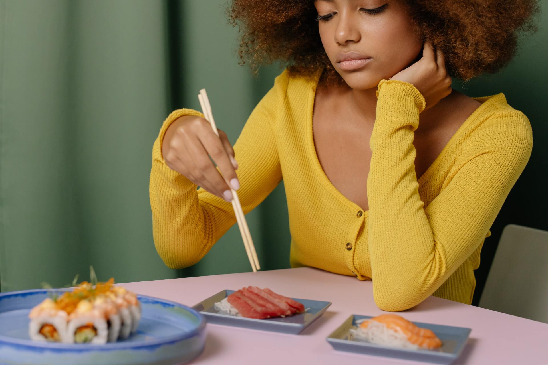 A woman is sitting at a table eating sushi with chopsticks.