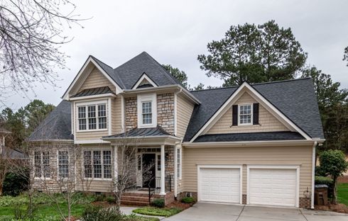A two-story suburban house with tan siding, stone accents, a dark shingled roof, and an attached two-car garage.