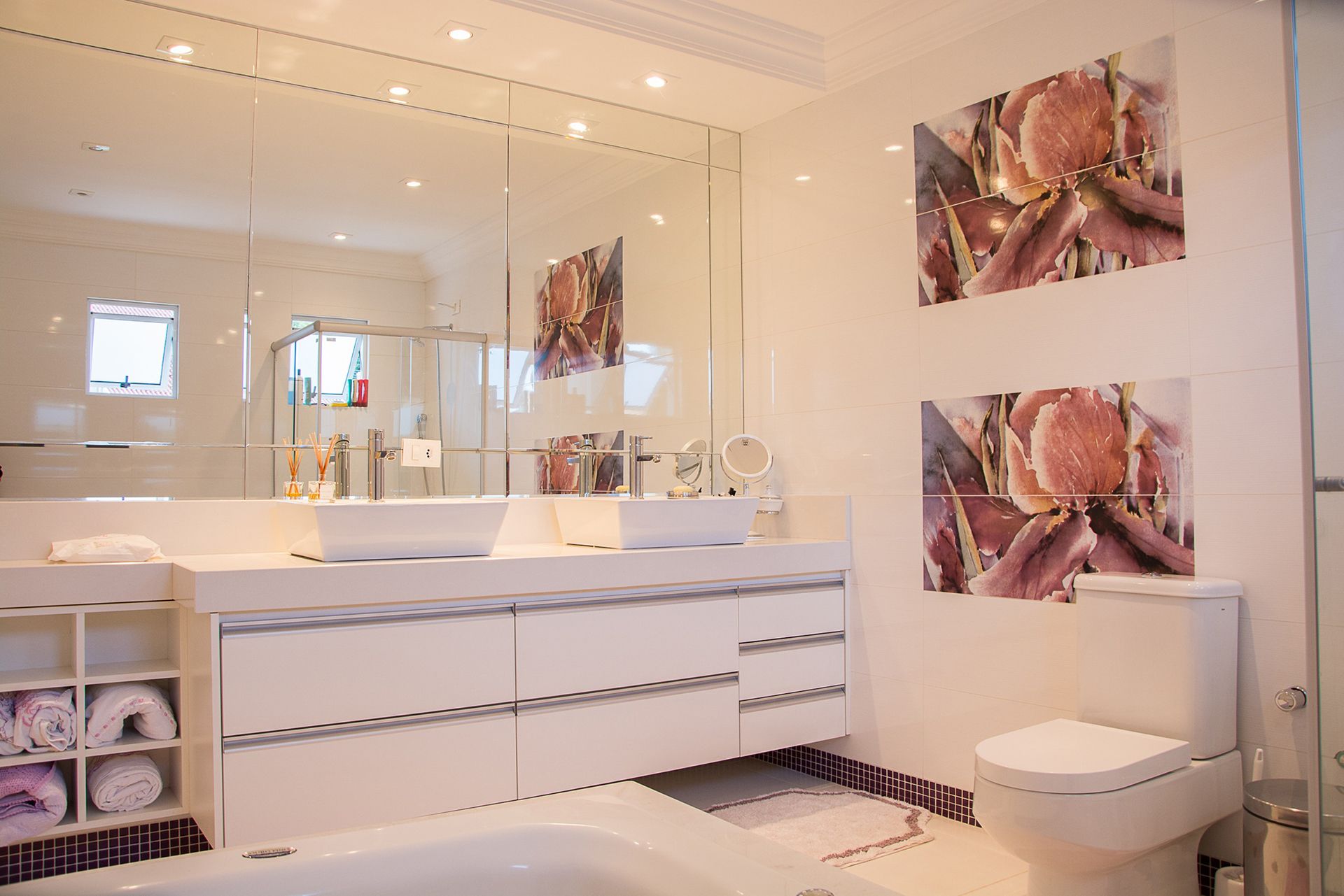 A bright, modern white bathroom with double sinks, large mirrors, a toilet, and floral wall art.