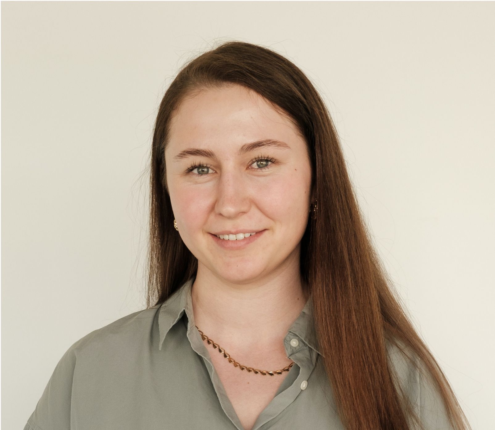A young woman with long brown hair smiles at the camera, wearing a gray button-down shirt, against a plain white background.