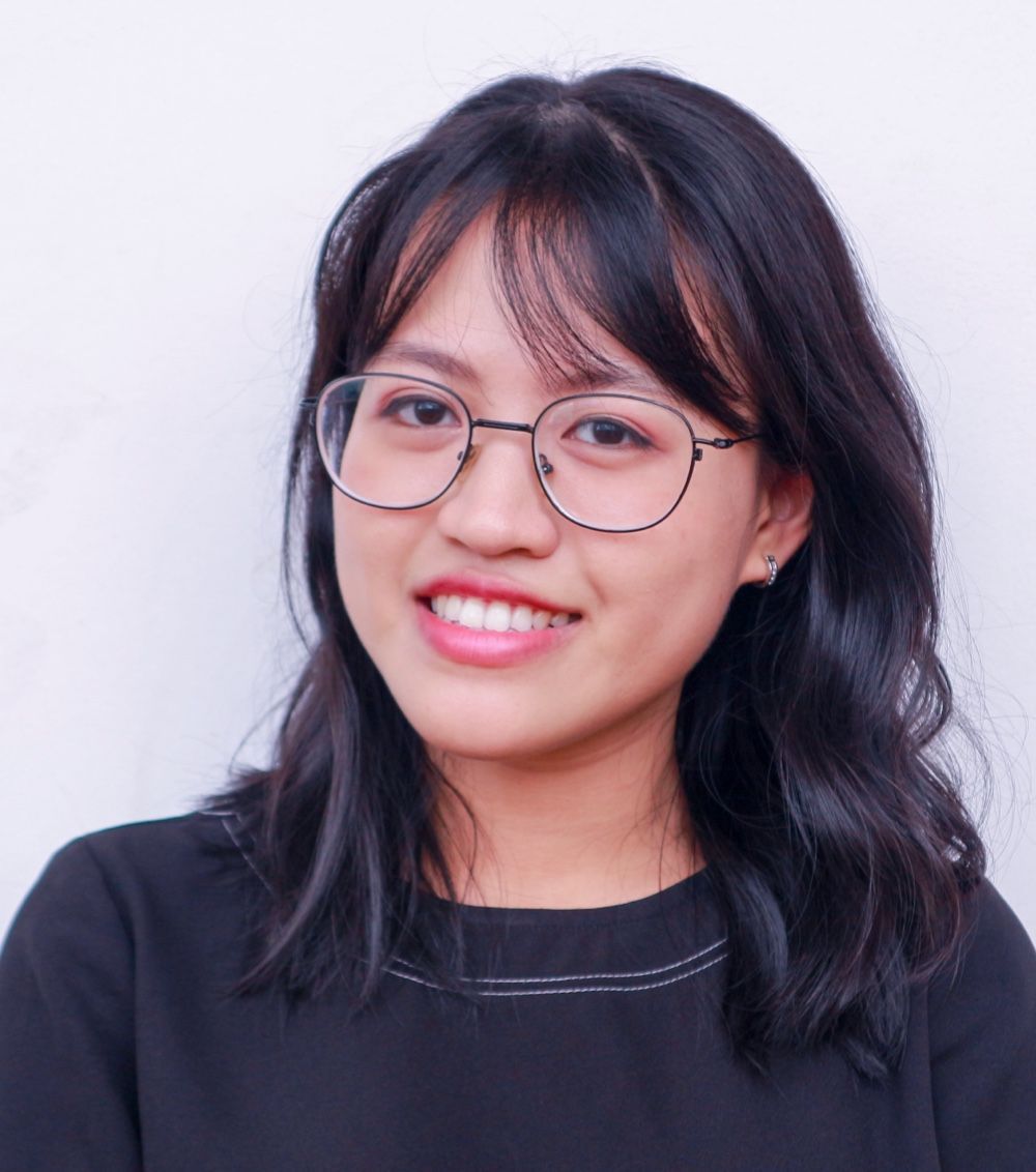 A smiling person wearing glasses and a black top against a plain, light-colored background.
