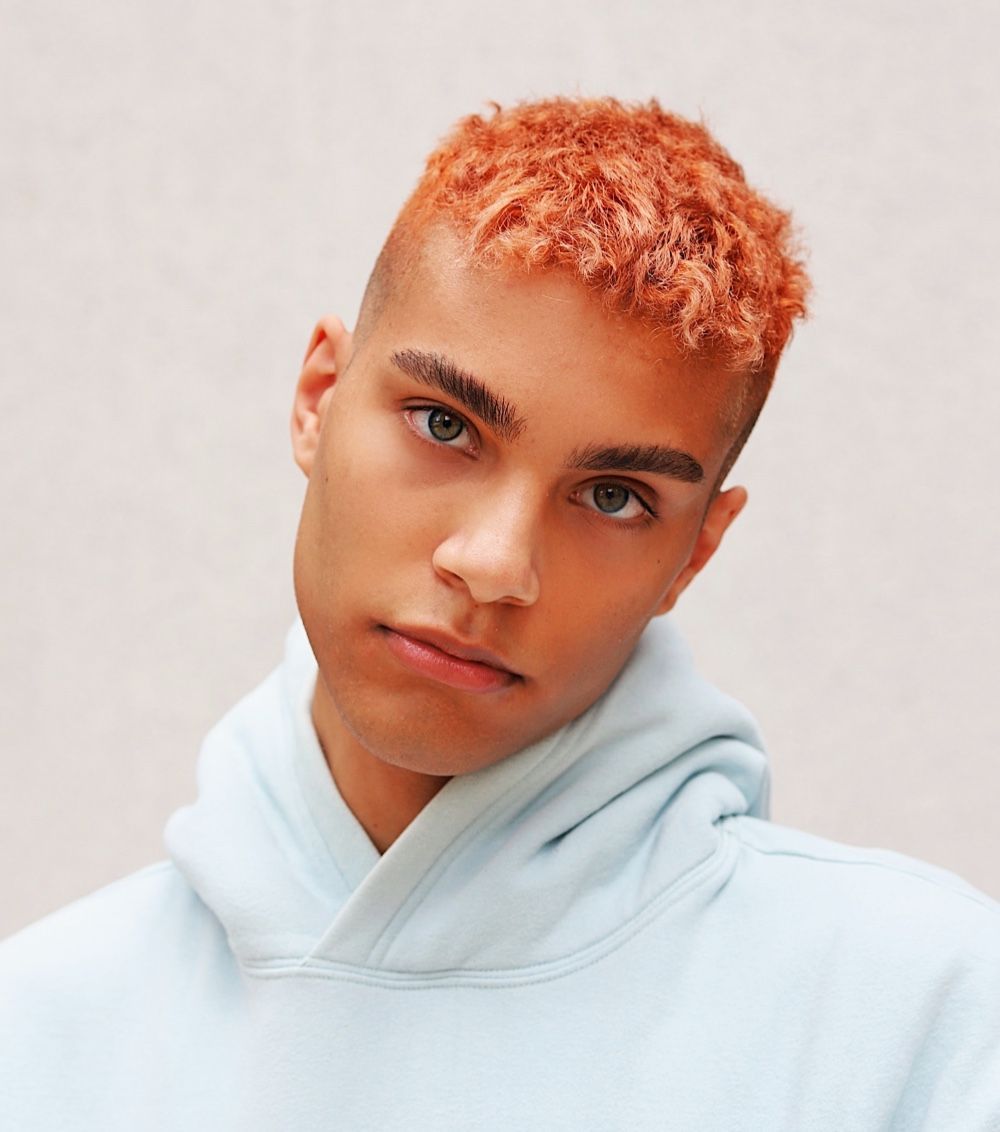 A person with short, curly, ginger-colored hair and a serious expression, wearing a light blue hooded sweatshirt.