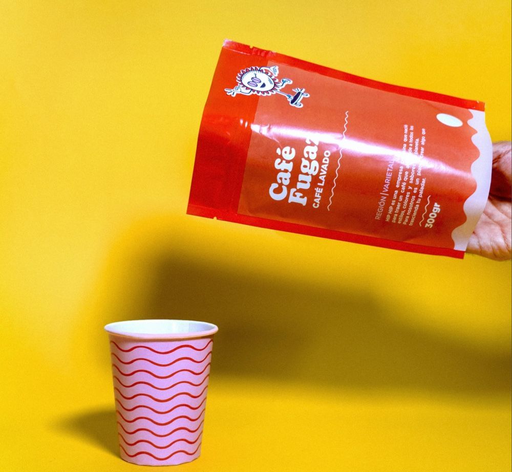 A hand holds an orange bag of Café Fugaz coffee beans over a small pink cup with a wavy pattern against a yellow background.