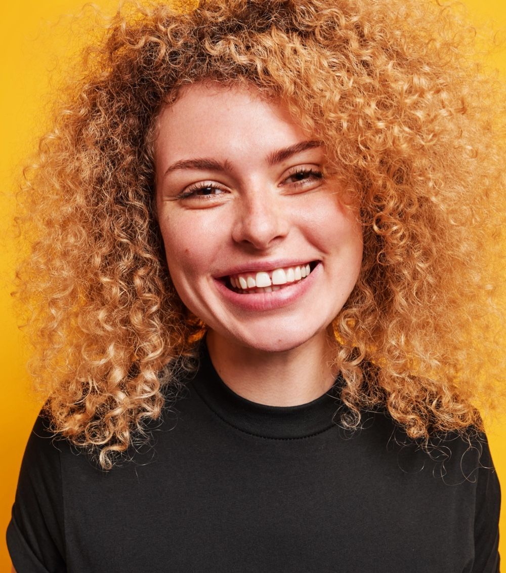 A smiling person with voluminous, curly, light-brown hair, wearing a black shirt against a plain yellow background.