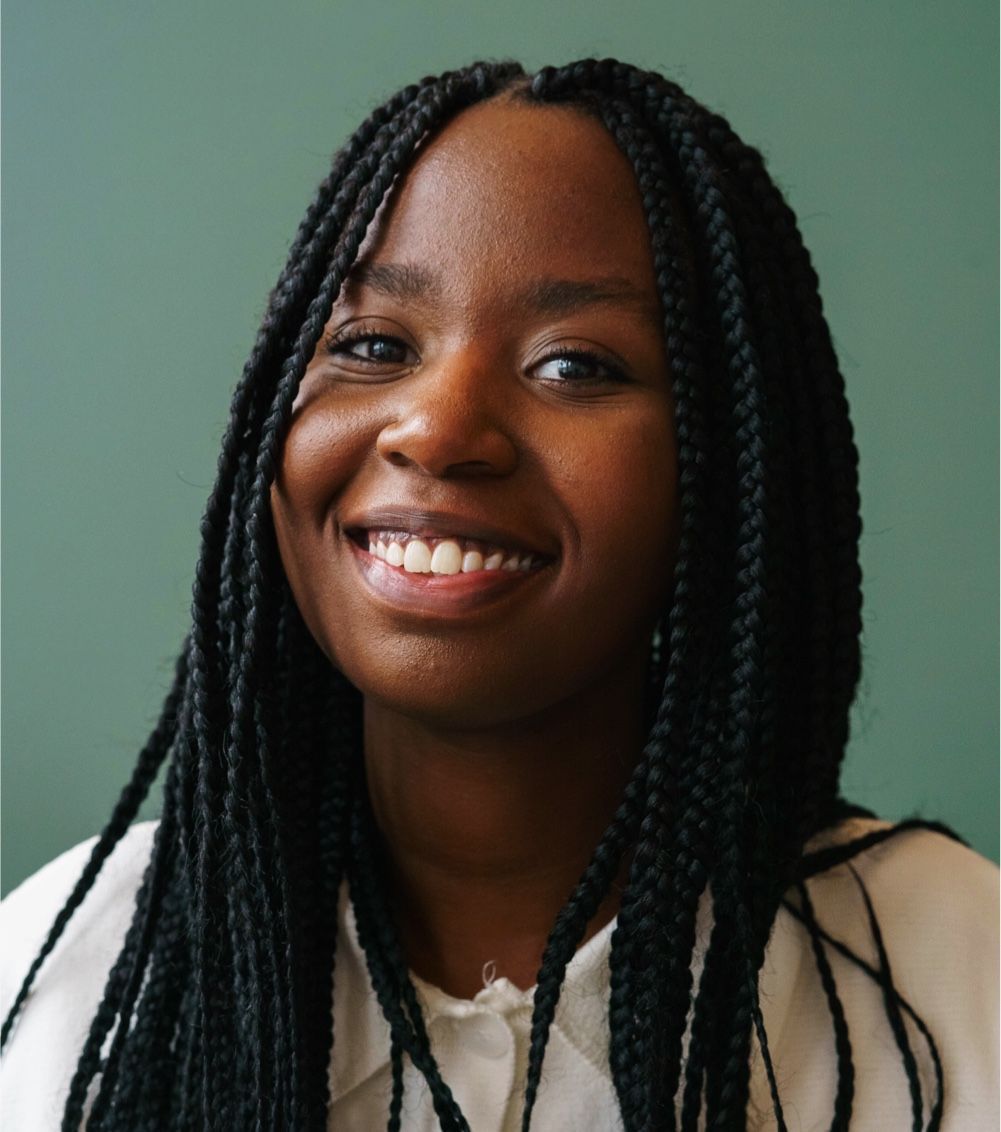 A person with braided hair smiling warmly against a solid sage green background.
